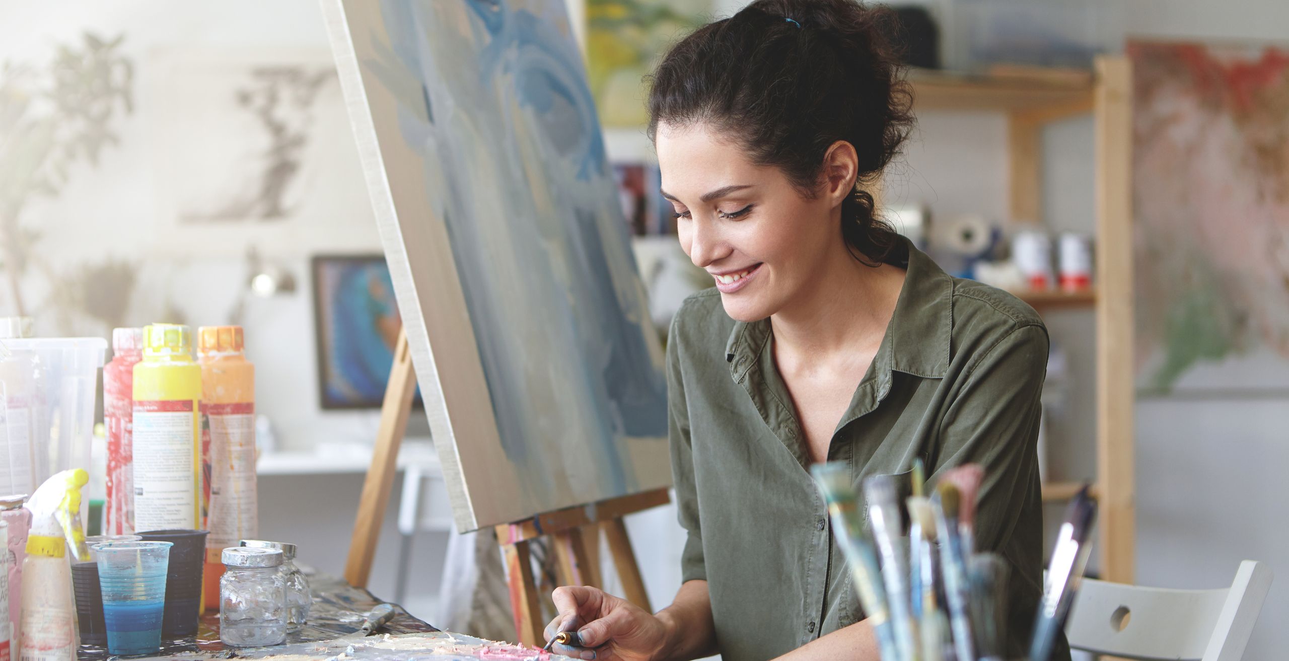 An artist smiling as she paints at her easel in a sunlit studio